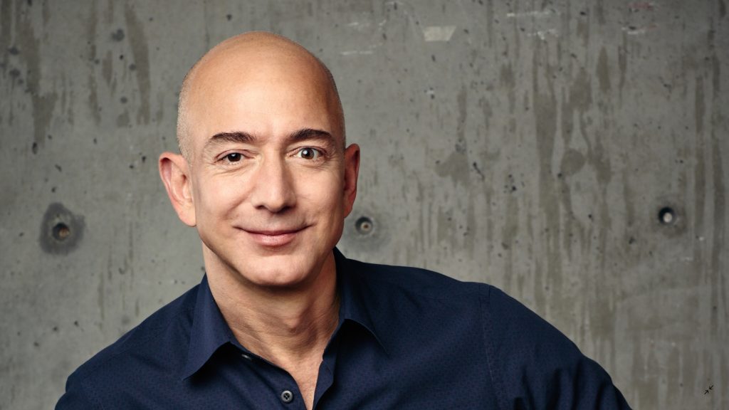 Jeff Bezos sorri enquanto posa em frente a uma parede cinza